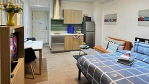 Una habitación de hotel compacta con kitchenette, una cama con colcha estampada en azul y blanco, una pequeña mesa de comedor y un televisor.