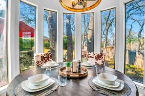 A dining area with a round table set for four, large windows, and a view of trees and a red building outside. - Spacious Gem, Hot Tub, 10 Min to AT&T & DFW | JCE (Grand Prairie)