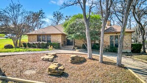 A residential property with a brick house, a landscaped yard with rocks, and mature trees. - Spacious Gem, Hot Tub, 10 Min to AT&T & DFW | JCE (Grand Prairie)