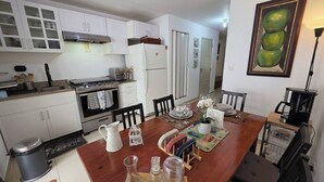 Een kleine keuken met witte kastjes, een fornuis en een koelkast. Er is een eettafel met stoelen en een koffiezetapparaat.