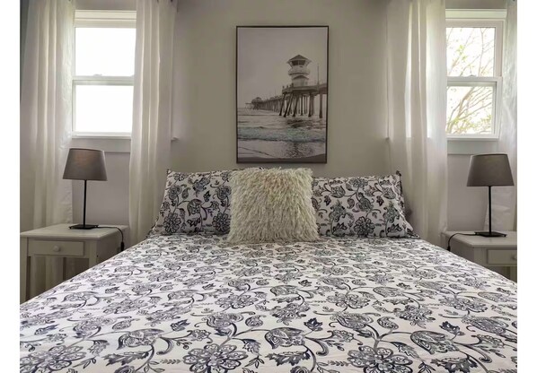 Un dormitorio con una cama con colcha de estampado floral, dos lámparas de noche, un almohadón blanco mullido y un cuadro con una imagen de un muelle en la pared.