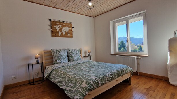 A bedroom with a wooden bed, a world map wall art, a window with a view of hills, and a radiator. - The House of Mountains (Le Thillot)