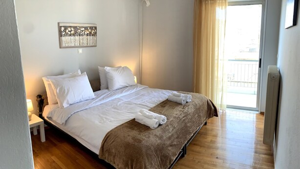 A bedroom with a bed, a nightstand, a wall-mounted light fixture, and a view of the outside through a window with curtains. - The Temple of Comfort (Athina)