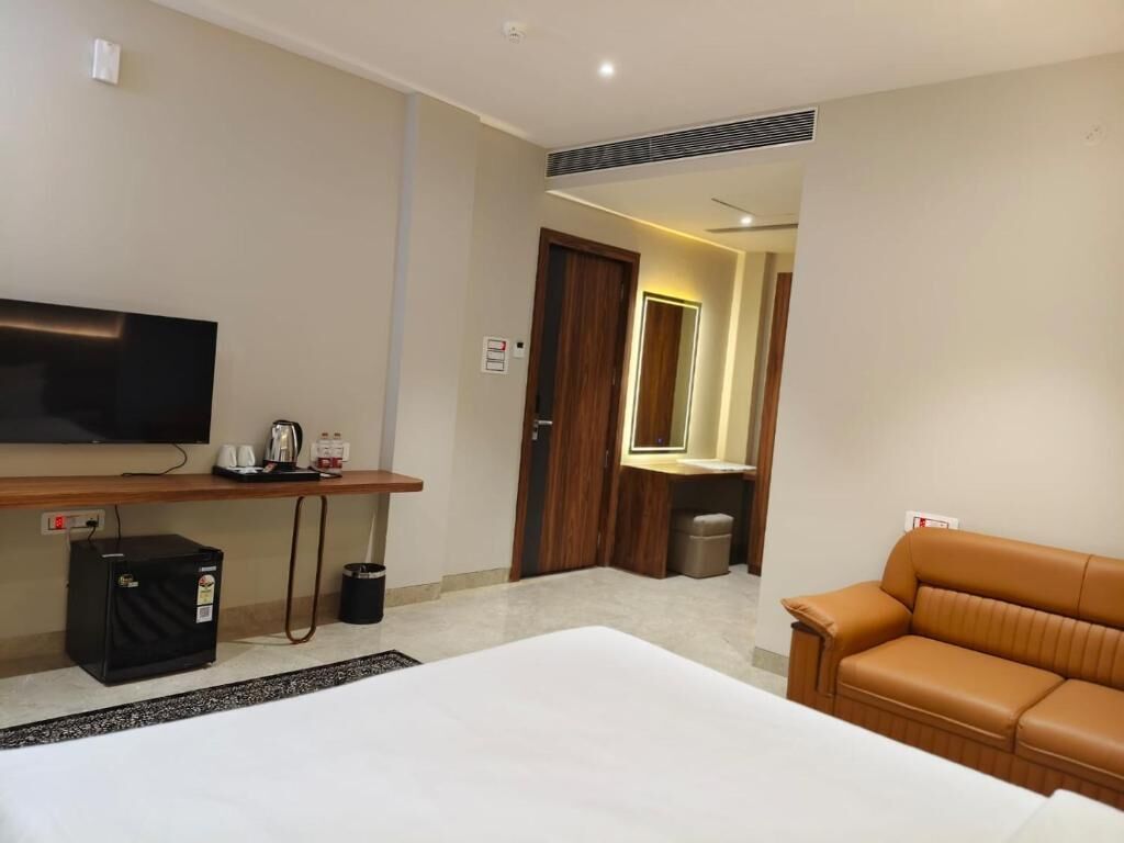 A hotel room with a bed, a television, a coffee machine, a small refrigerator, a trash bin, a brown leather sofa, and a mirror.