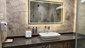 A bathroom with a marble wall, a large mirror, a white sink, and a glass shower enclosure. - THE CROWN TOWN BIDAR (Bidar)