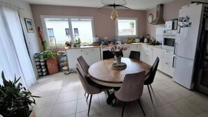 A modern kitchen with white cabinets, a dining table with chairs, and a refrigerator. - Single-storey seaside house (Santec)