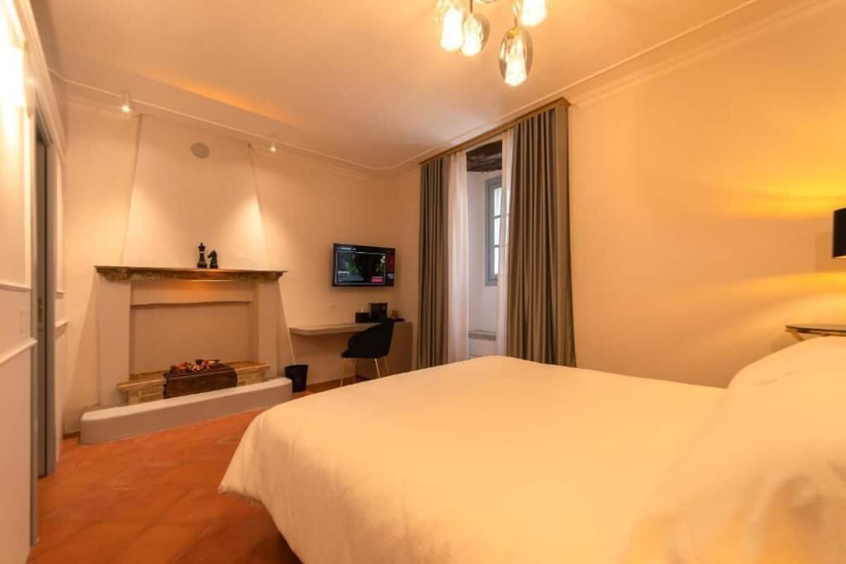 A hotel room with a large bed, a desk with a chair, a television, and a fireplace.