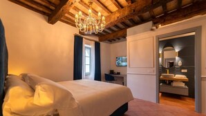 A bedroom with a large bed, wooden ceiling beams, a chandelier, a desk, and a bathroom with a mirror and sink. - Maison Ventiquattro Suite & Spa (Talamello)