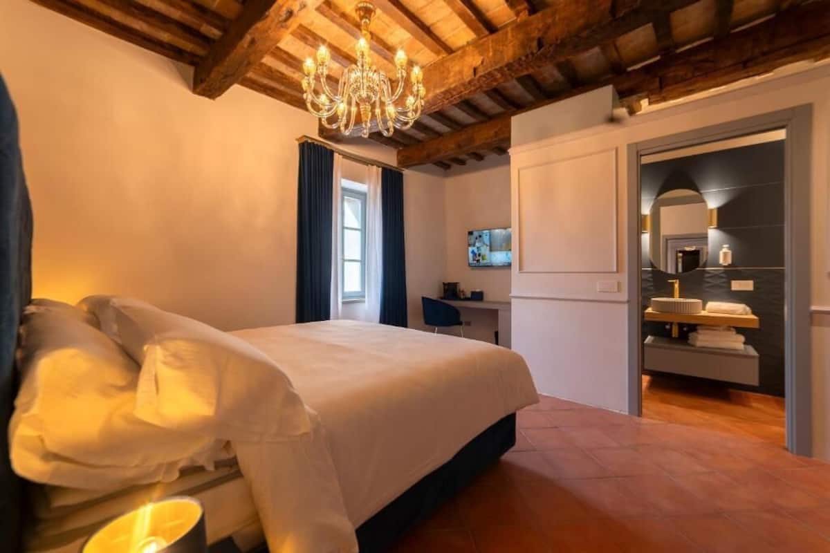 A bedroom with a large bed, wooden ceiling beams, a chandelier, a desk, and a bathroom with a mirror and sink.