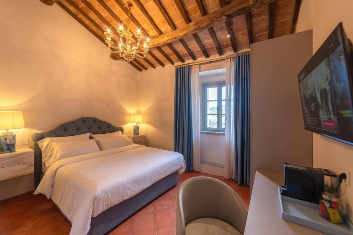 A bedroom with a bed, nightstands, a desk, a chair, a chandelier, and a window with curtains.