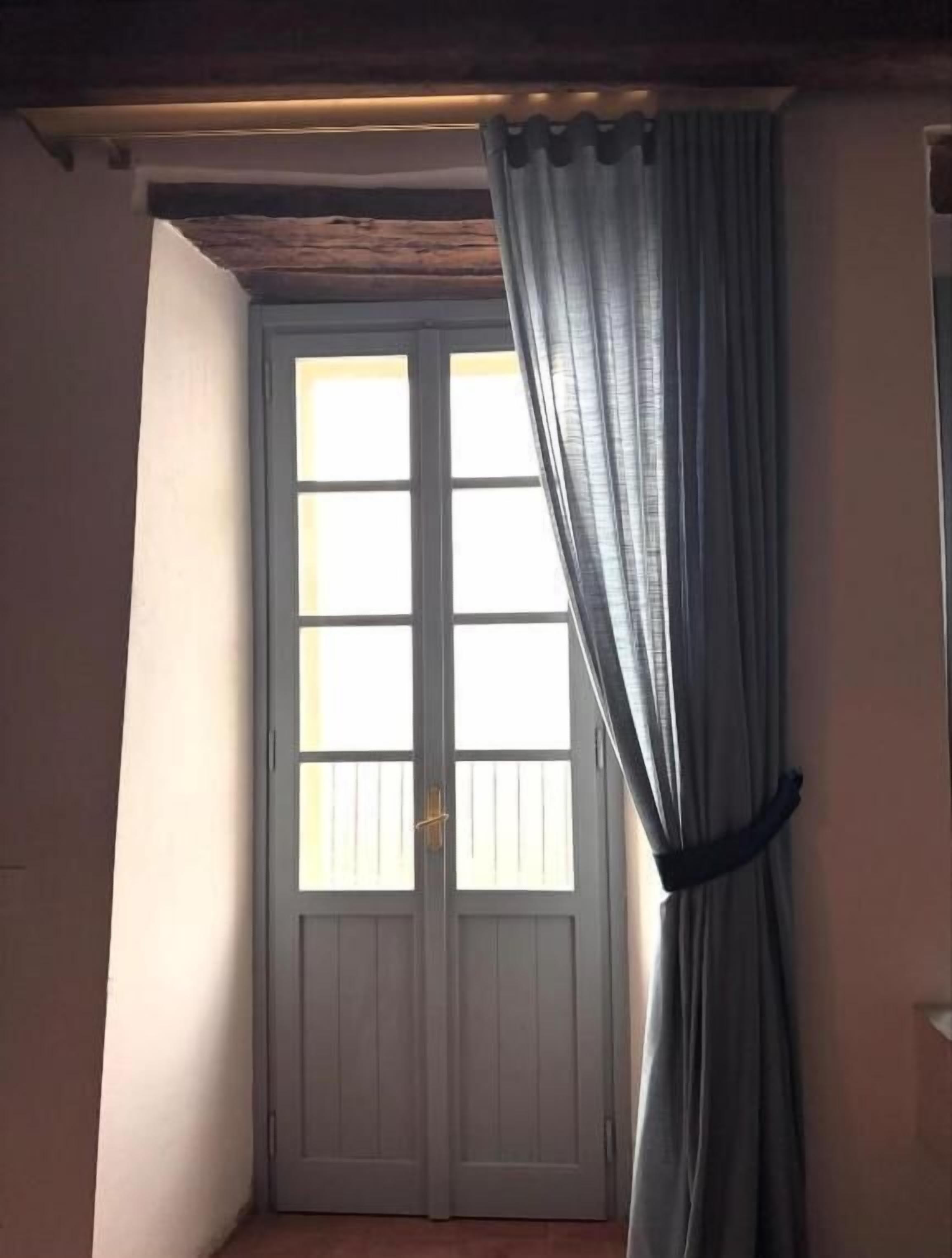 Ein Zimmer mit doppeltürigem Zugang, einem Fenster mit Vorhang und einem Holzbalken an der Decke.
