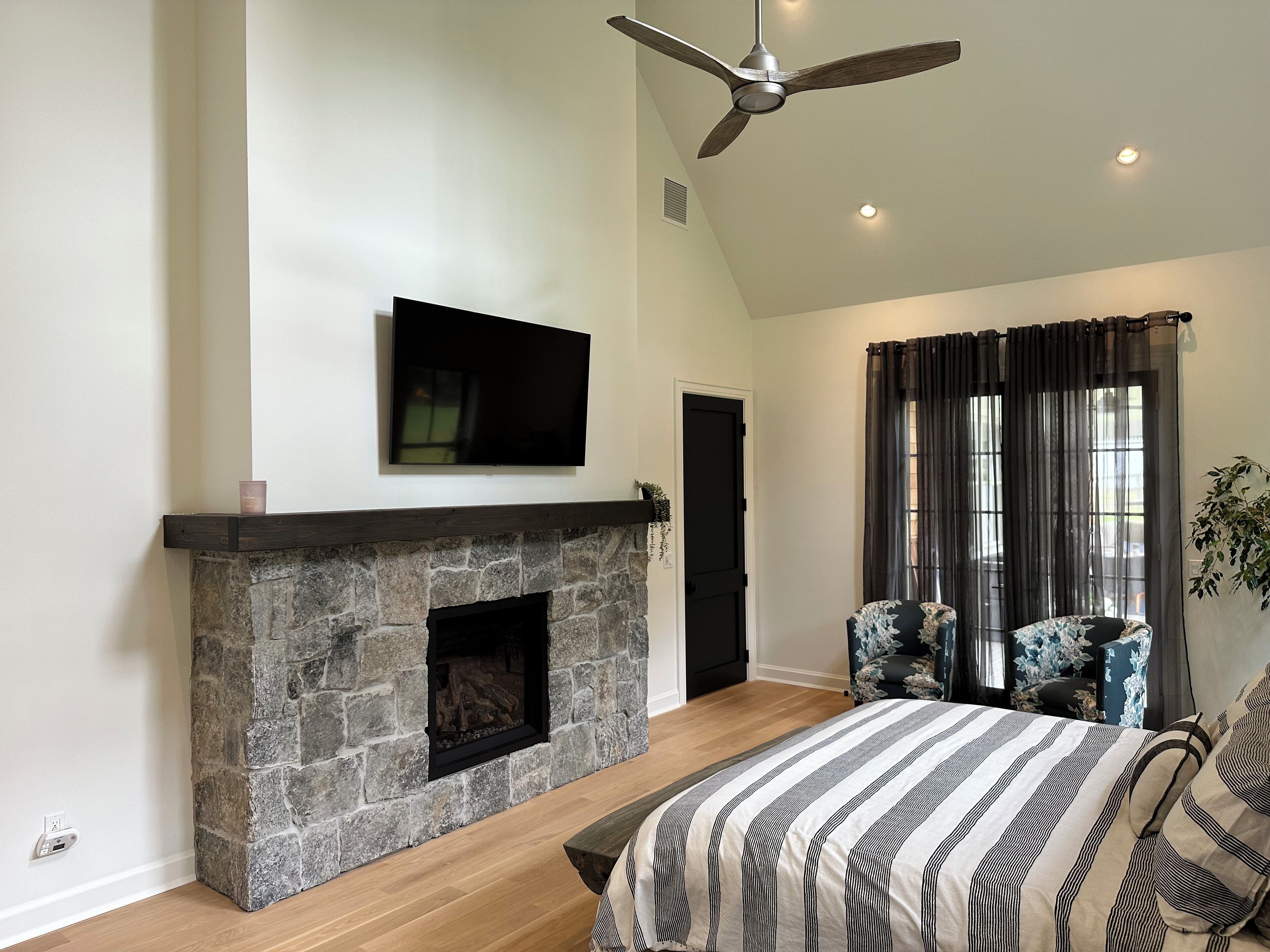 A bedroom with a stone fireplace, a ceiling fan, a bed with striped bedding, and a large window with curtains.
