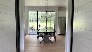Un pasillo con un banco de madera, una mesa de comedor con sillas, una araña y vistas al exterior a través de amplios ventanales.