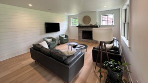 Una sala de estar con chimenea, un sofá, dos sillones, una mesa de centro y un televisor montado en la pared.