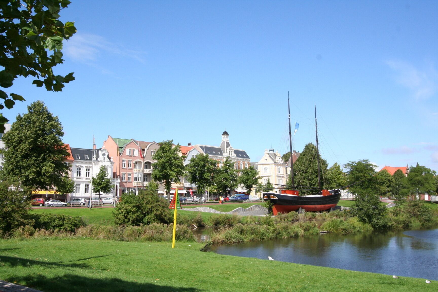 A riverside view with a historic building, a sailboat, and a grassy area.