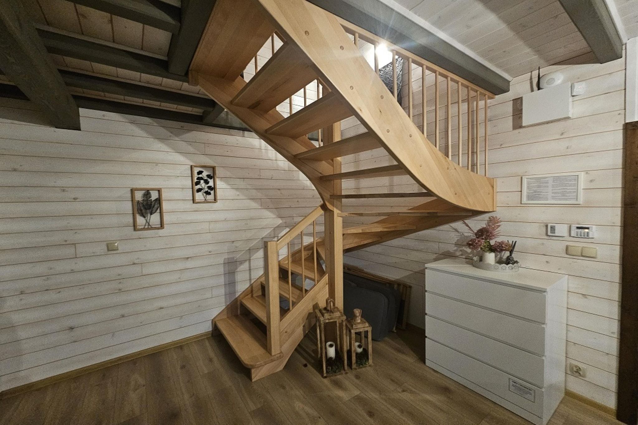 Una escalera en espiral de madera en una habitación con paredes de madera blanca, un armario blanco y cuadros enmarcados.