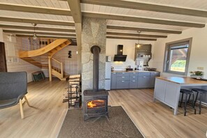 Un interior acogedor con estufa de leña, una cocina con armarios grises, una escalera de madera y un ventanal grande.