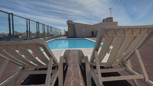 Un área de piscina en la azotea con dos sillones blancos mirando a la piscina, un cielo azul despejado y un sol brillando tras las nubes.