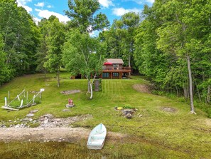 A cabin with a deck, surrounded by a grassy area and trees, near a body of water with a boat.