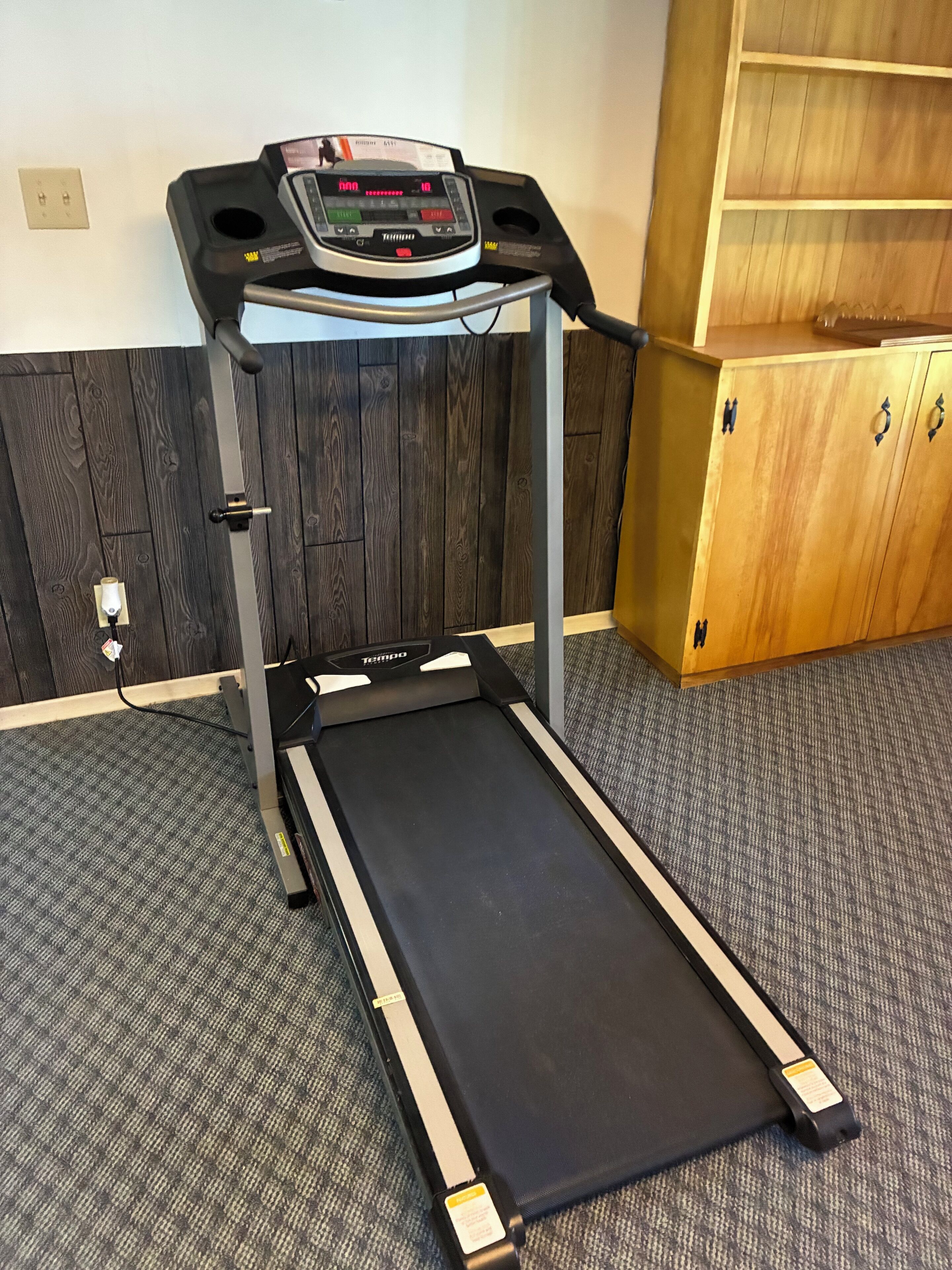 A treadmill in a room with a wooden cabinet and a carpeted floor.