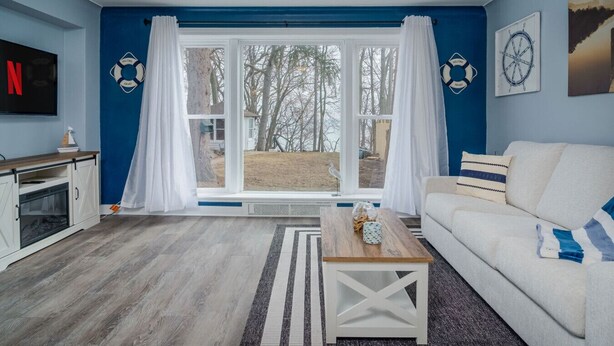 A living room with a large window, a white sofa, a wooden coffee table, and a TV mounted on the wall. - Mineola Extended-Stay Furnished 30-Day+ Cottage (Fox Lake)