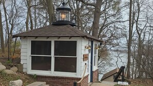A small, white, single-story building with a brick base and a dark roof. It features a large window and a small porch area. - Mineola Extended-Stay Furnished 30-Day+ Cottage (Fox Lake)