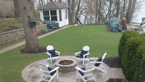 A patio area with four Adirondack chairs around a circular fire pit, a brick pathway, and a view of a house and trees. - Mineola Extended-Stay Furnished 30-Day+ Cottage (Fox Lake)