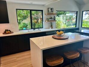 A modern kitchen with a central island, bar stools, a large window with a view of greenery, and a built-in shelf with decorative items. - Casa Sharman in Solana Beach welcomes you to live with ease and joy. (Solana Beach)