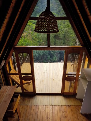 An attic room with a wooden floor, a hanging light fixture, and a view of a green landscape through a large window. - Ecolodge Ix Cacao  (San Carlos)