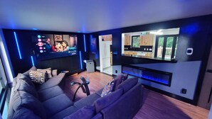 Un salon moderne doté d’un grand téléviseur à écran plat, d’un canapé et d’un coin cuisine visible à travers une porte ouverte.