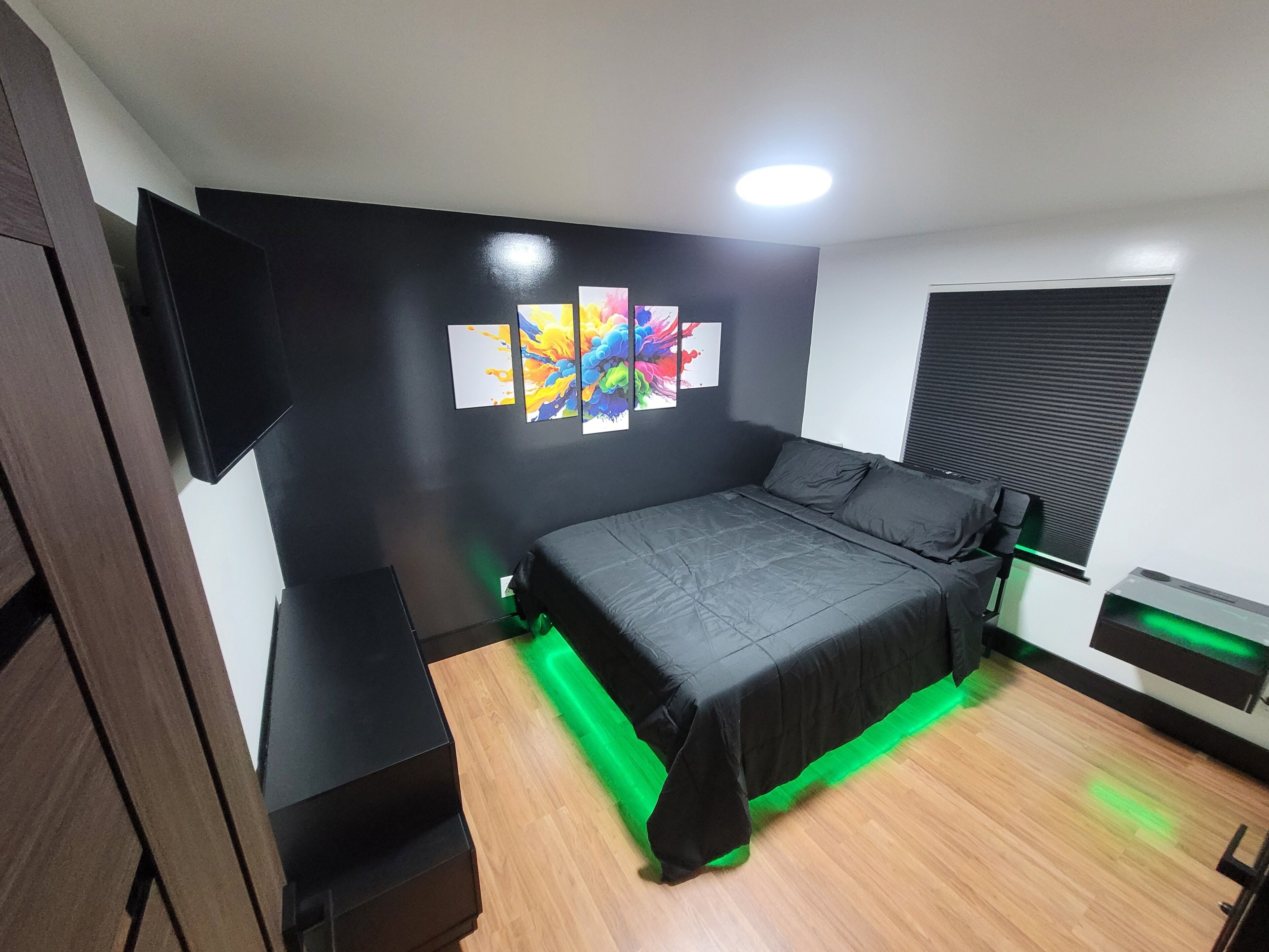 A bedroom with a bed, a TV mounted on the wall, and a window with blinds.