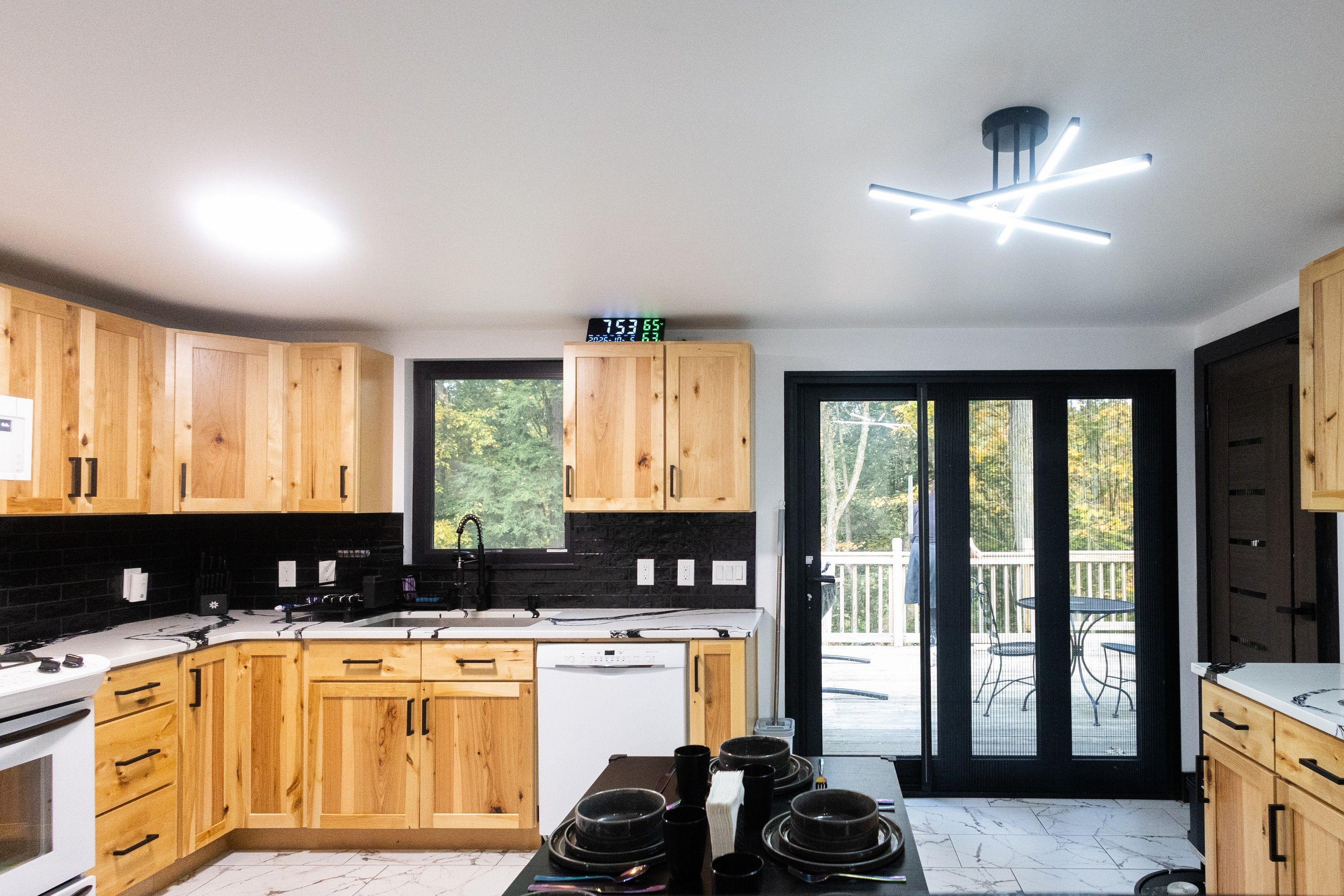 A modern kitchen with wooden cabinets, a digital clock, and a view of the outdoors through a sliding glass door.
