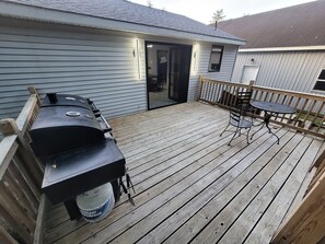 Une terrasse en bois avec un barbecue, une table et des chaises, et une porte-fenêtre coulissante donnant sur un espace abrité.