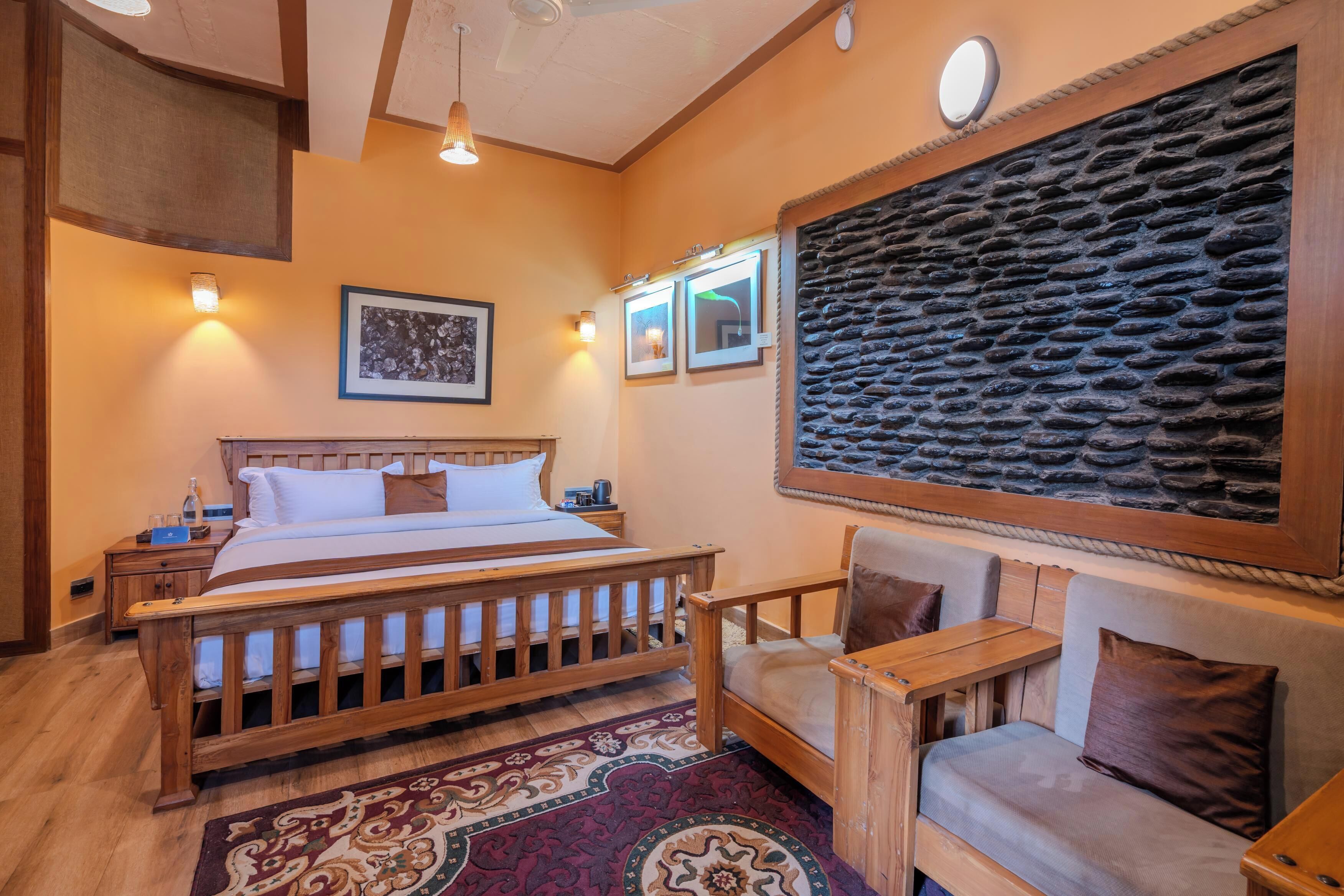 Un dormitorio con cama de madera, una mesita de noche, una silla, alfombra estampada y pared de piedra.