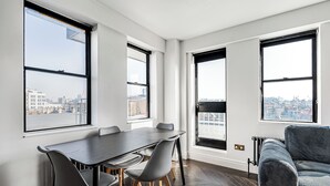 Une pièce moderne comprenant une table à manger, des chaises et un canapé. De grandes fenêtres offrent une vue sur la ville.