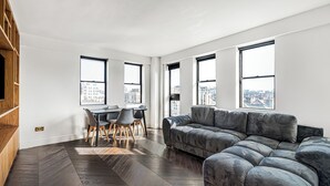 Un salon moderne avec un canapé d’angle, un coin repas comprenant une table et des chaises, et de grandes fenêtres offrant une vue sur la ville.