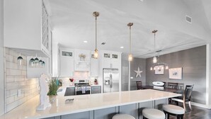 A modern kitchen with a central island, bar stools, pendant lighting, and a dining area with a starfish decoration. - Seashell Shack on Shrimp, Sleeps 11 - 3 bedroom 2.5 Bath - Great View (Crystal Beach)