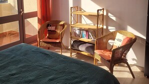 A room with a bed, two wicker chairs, a bookshelf, and a painting of stones.