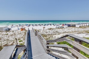 En strand med en träpromenad som leder ner till stranden, med strandparasoller och solstolar.