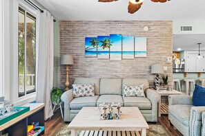 A living room with a brick wall, a sofa set, a coffee table with toys, and a view of the ocean through a window. - 🏖️Lagoon Pool | Hot Tub | Walk to Baytowne Wharf | Market Street Inn 248☀️ (Miramar Beach)