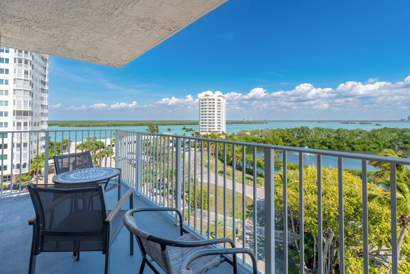 Un balcon avec deux chaises et une table, offrant une vue sur la côte, avec un ciel dégagé et un bâtiment visible au loin.