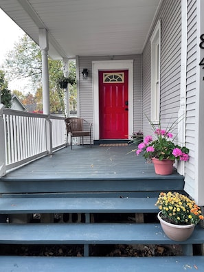 Un porche avec une porte rouge, une chaise en osier et des plantes en pot.