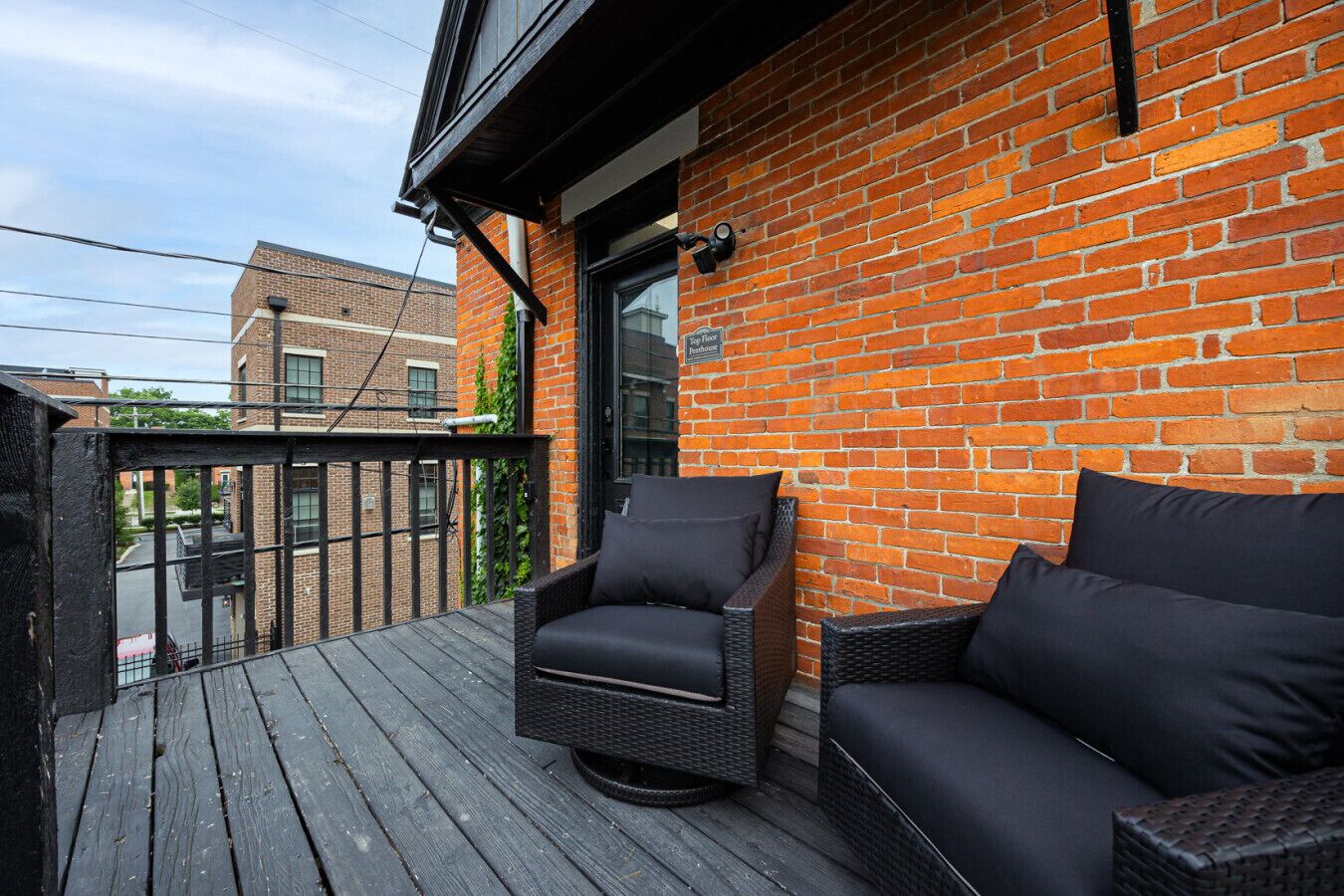 A balcony with a wooden deck, two wicker sofas with dark cushions, and a brick wall.