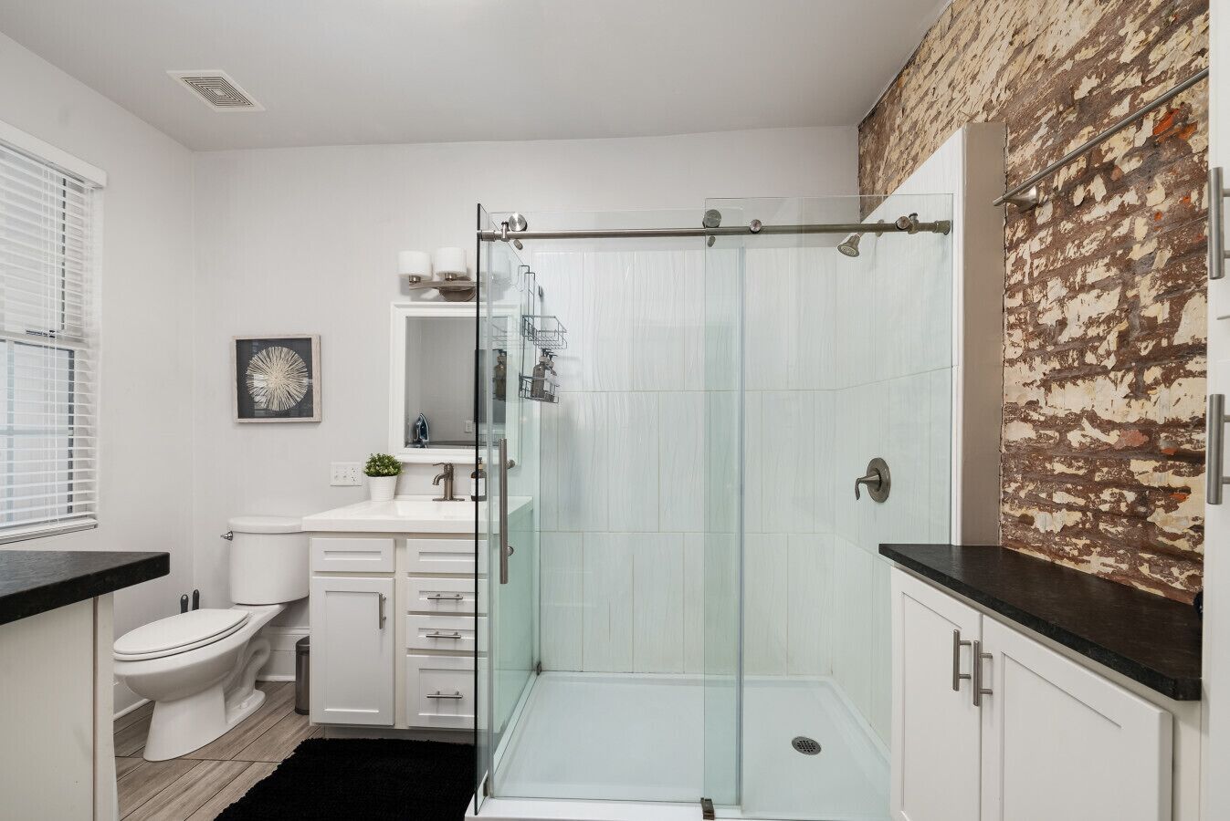 Un baño moderno con cabina de ducha de vidrio, muebles blancos y una pared de ladrillo decorativa.