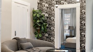 Een kamer met een grijze bank, een grote groene plant en een zwart geweven poef.