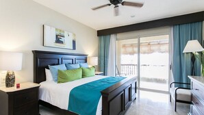 Quarto de hotel com cama, mesa de cabeceira, escrivaninha, cadeira, ventoinha no teto e uma janela grande com cortinas.
