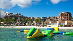 Um resort à beira-mar com águas azul-claras, brinquedos infláveis para a água e vários edifícios ao fundo.