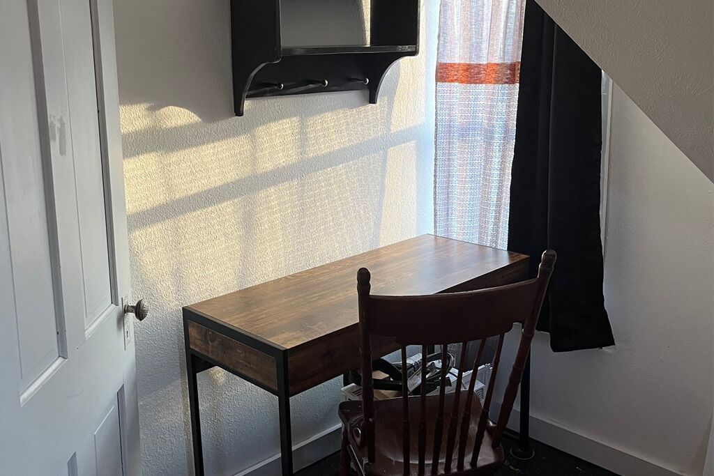 A room with a wooden desk, a chair, a window with curtains, and a mounted television.