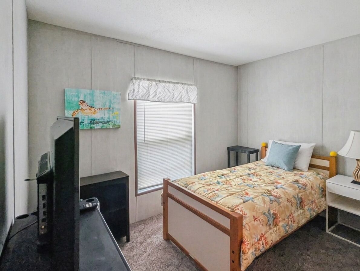 Quarto com cama, escrivaninha, cadeira, televisão, janela com persianas e um quadro na parede.
