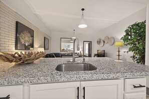 A kitchen with granite countertops, a stainless steel sink, white cabinetry, a hanging light fixture, and a dining area in the background.